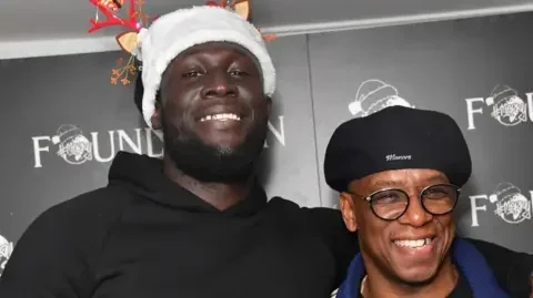 Getty Images Stormzy wears a black hoodie and a festive hat with antlers. He has his arm around Ian Wright, who is wearing black glasses and a black cap.