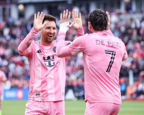Lionel Messi and Rodrigo De Paul in pink uniforms celebrate with a high-five on a field.